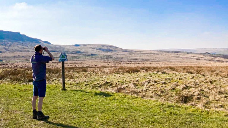 A volunteer uses binoculars to look over Marsden Moor on a sunny day